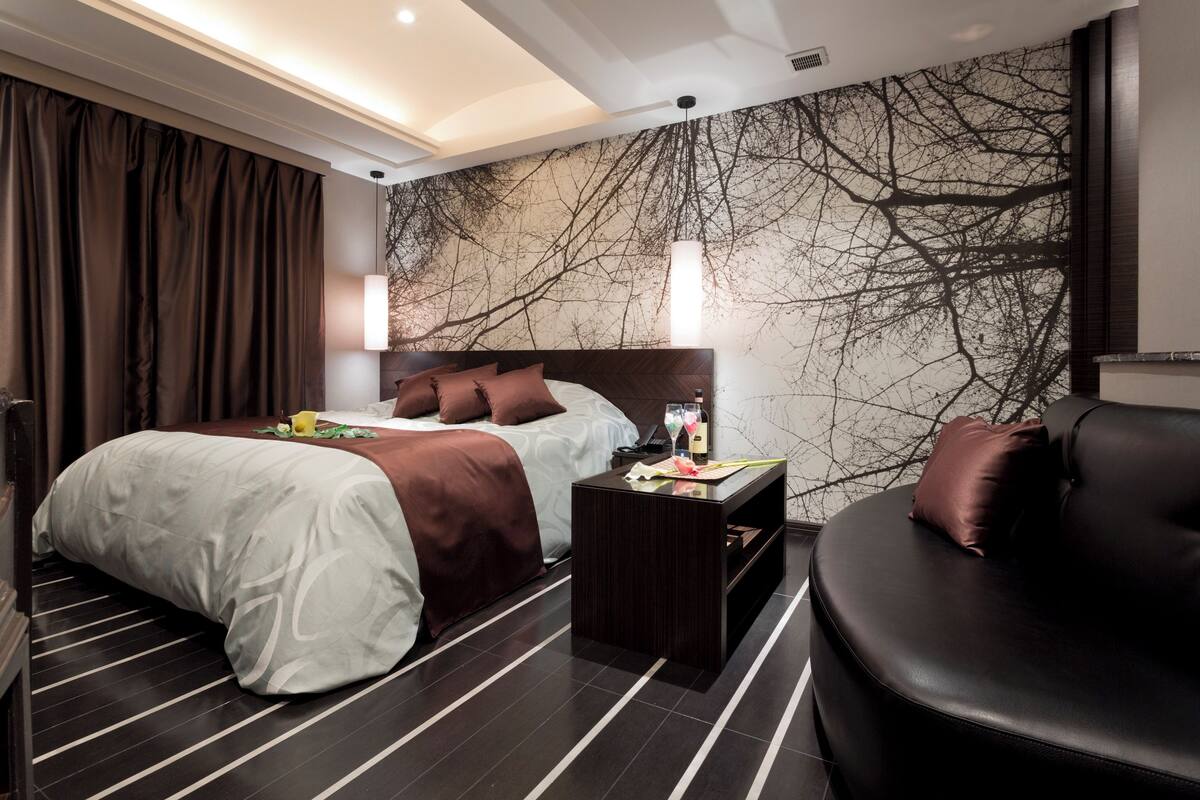 A modern hotel room with a large bed, a sofa, a nightstand, and a wall with a tree branch design.