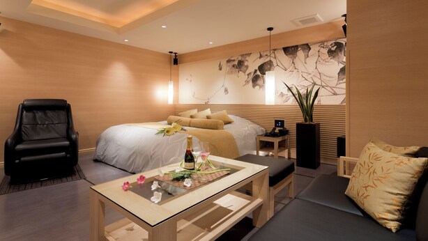 Una habitación de hotel moderna con cama, sofá, sillón, una mesita de cristal y una gran obra de arte en la pared.