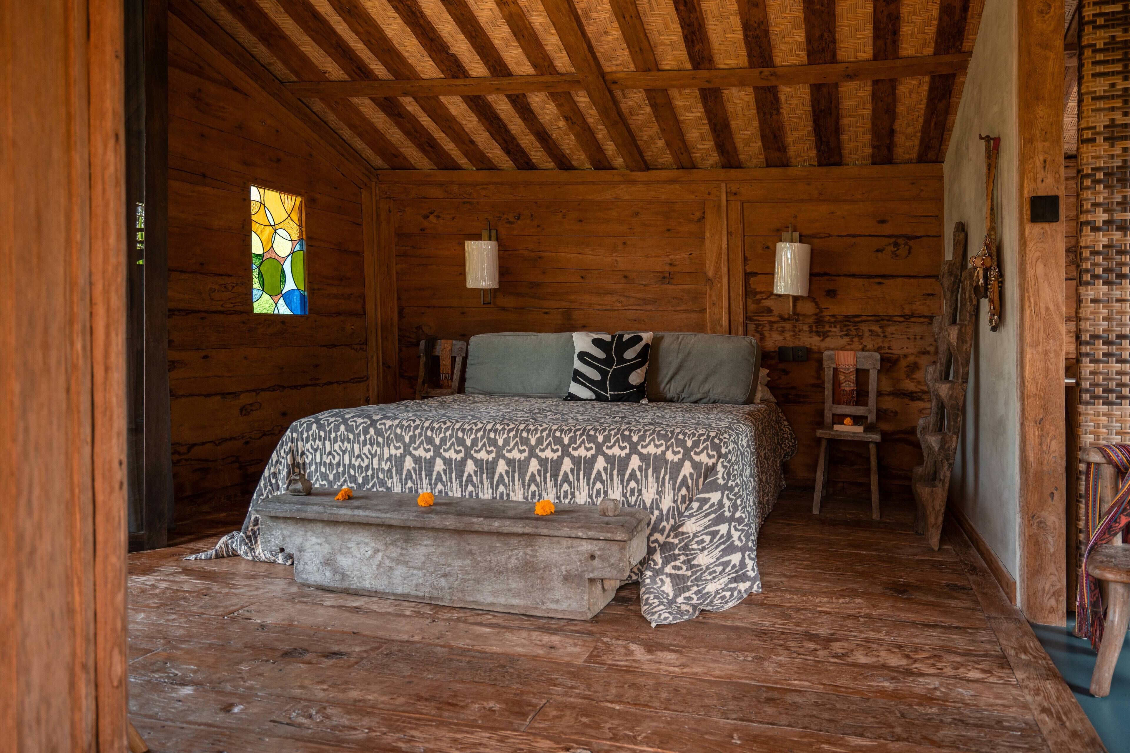 A wooden cabin bedroom with a bed, two wall sconces, a small stool, and a stained glass window.