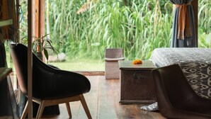 A room with a wooden ceiling, a bed with patterned bedding, a chair, a small table, and a view of lush greenery outside. - Desa Seni  (Baturiti)