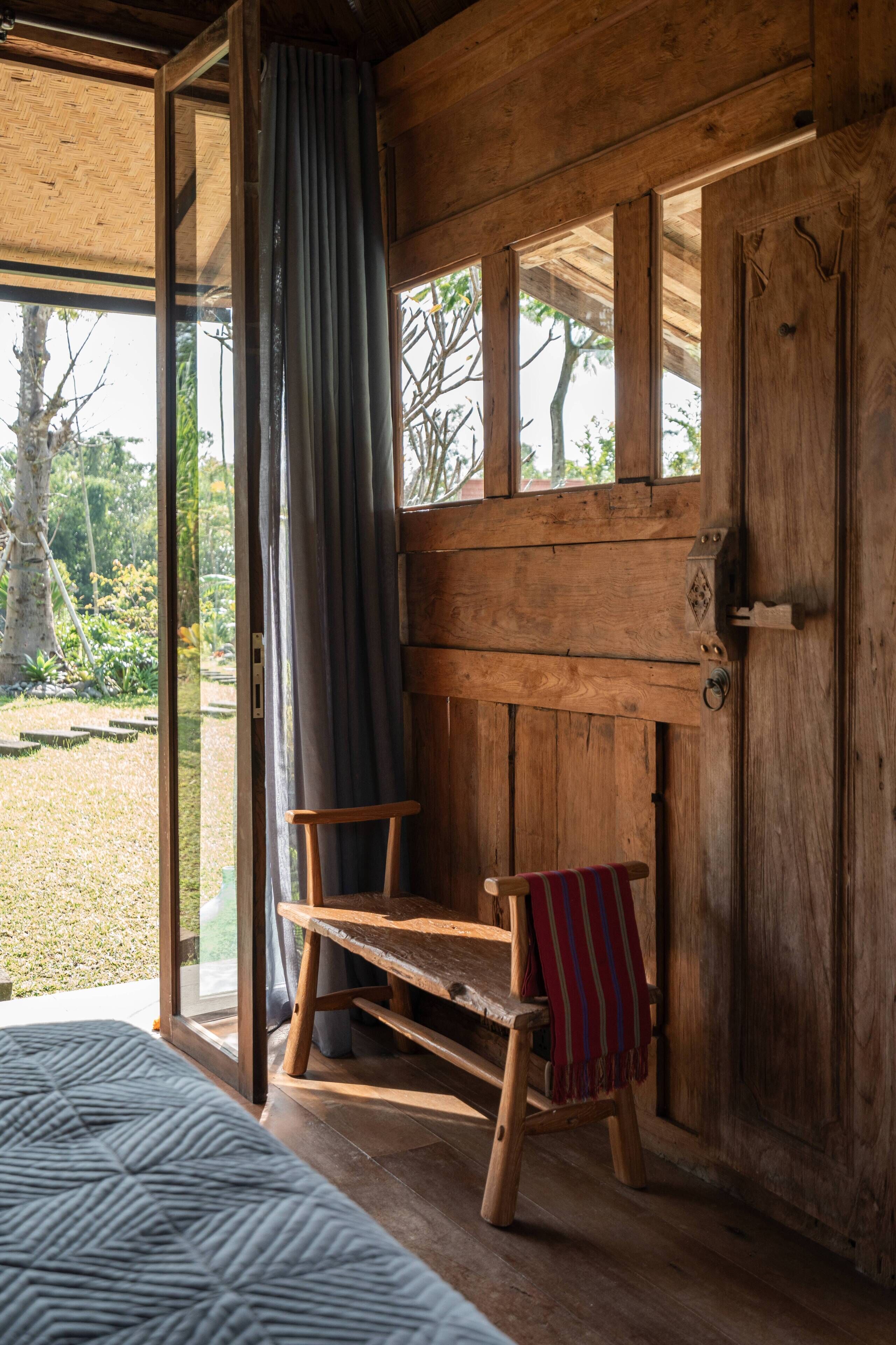 Une pièce de cabane en bois comprenant un lit, un banc avec une couverture à rayures, une fenêtre avec un rideau et une porte.