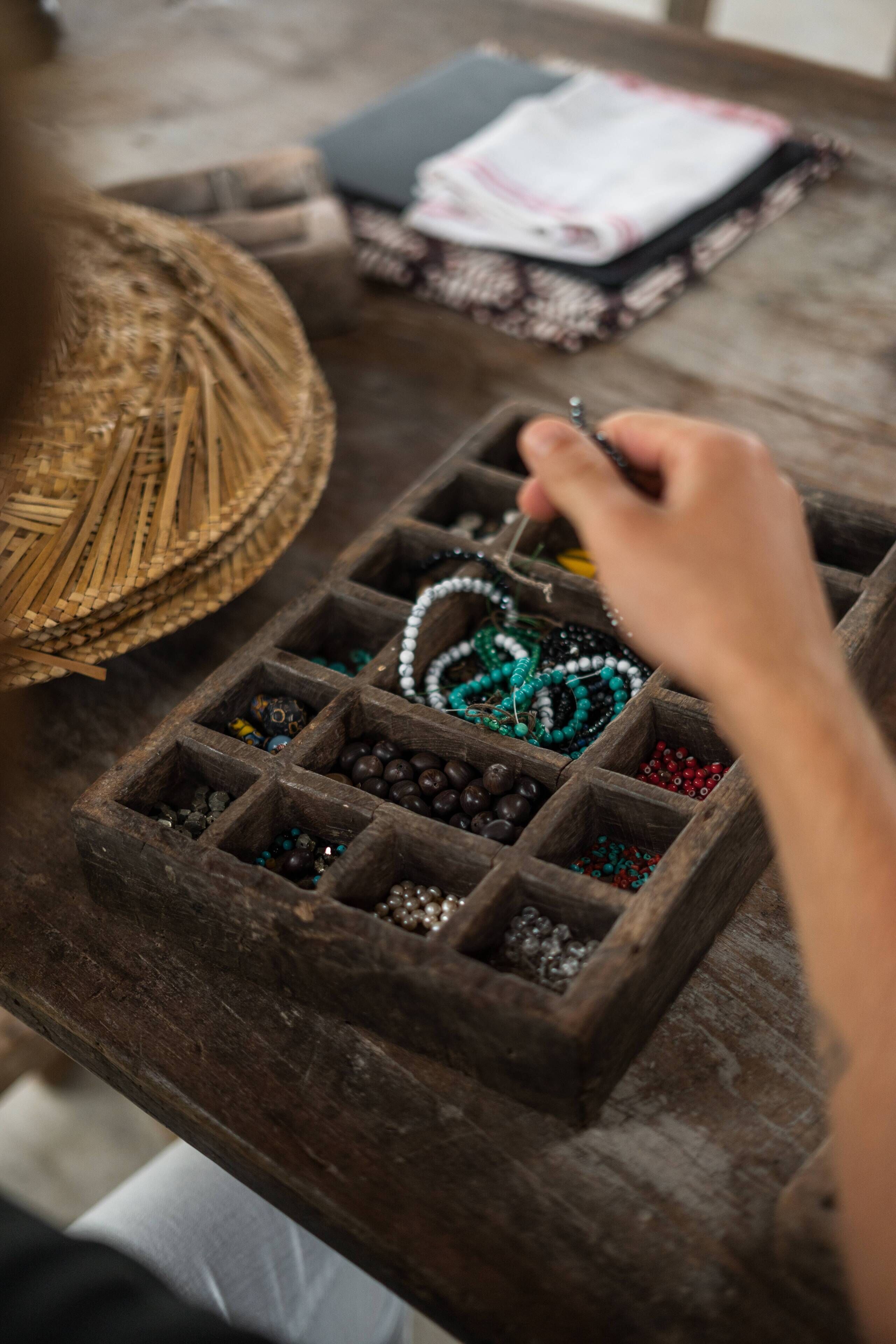 A wooden jewelry box with compartments containing various beads and a hand holding a string of beads.
