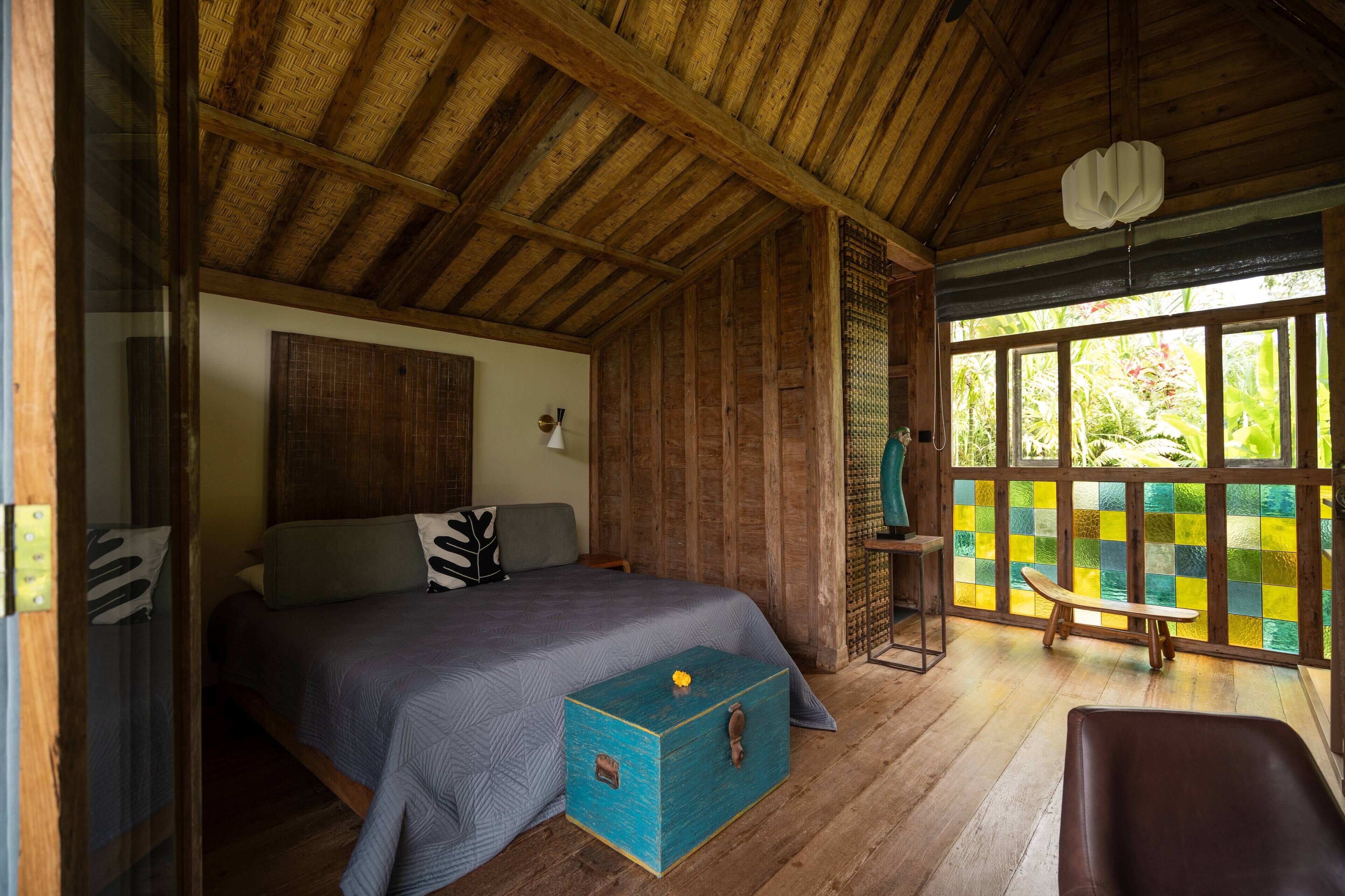Una habitación de madera con una cama, un cofre azul y un banco junto a una ventana con vistas al follaje.