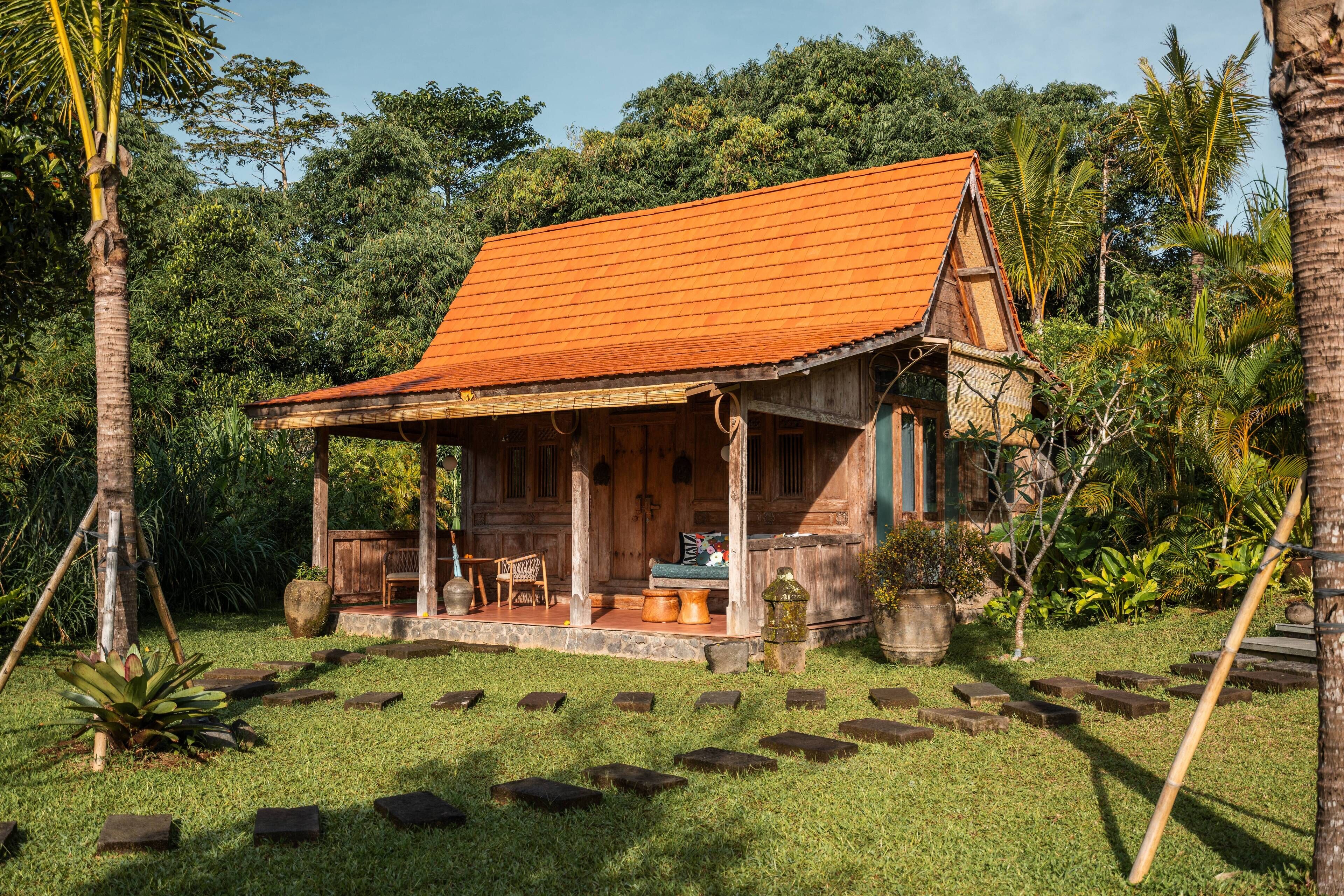 A wooden house with a tiled roof, surrounded by lush greenery and palm trees.