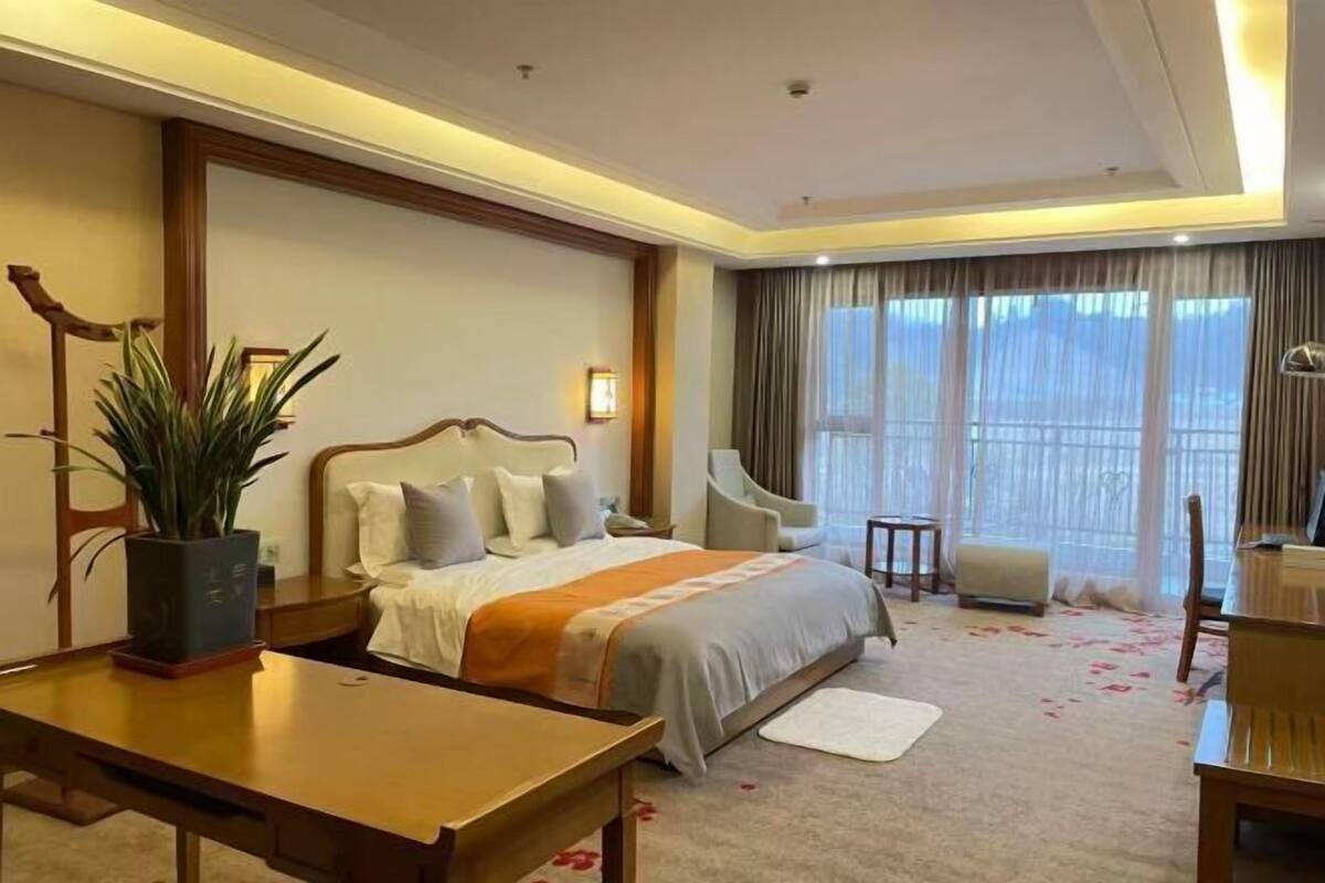 A hotel room with a bed, bedside table, chair, and a large window with curtains.