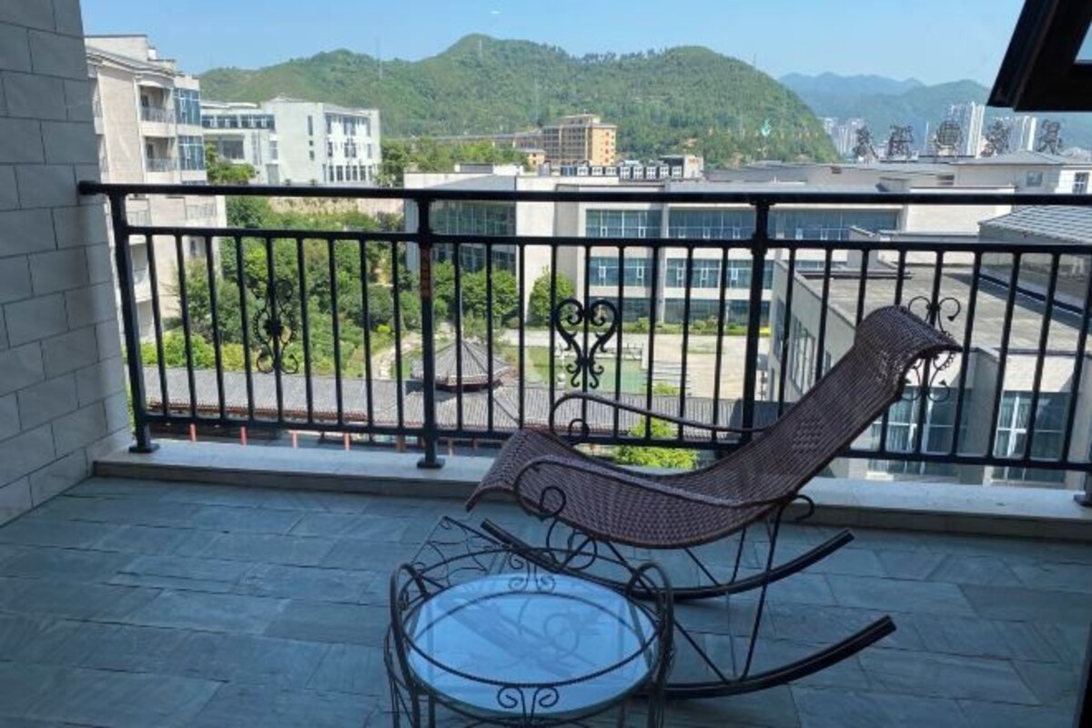 Un balcón con una silla de mimbre y una mesa de vidrio, con vistas a un paisaje urbano que incluye edificios y una colina.