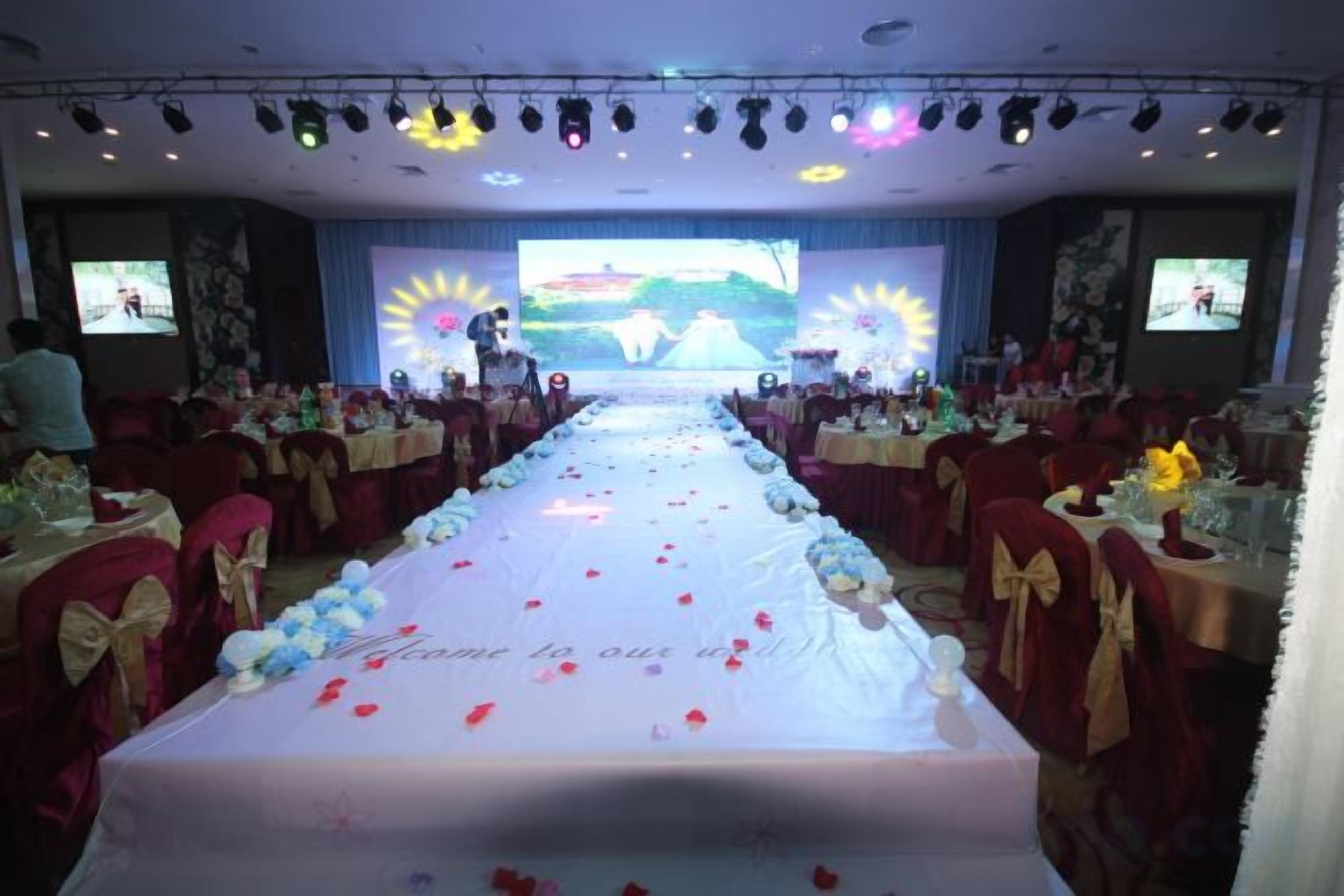 A banquet hall with a long table covered in a white tablecloth, red chairs, and a stage at the front.