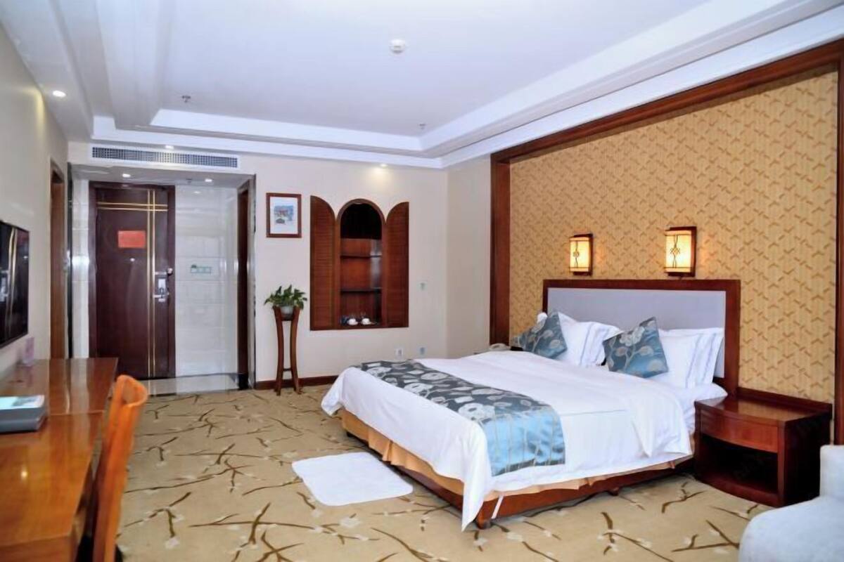 A hotel room with a bed, bedside table, lamp, and a desk with a chair.