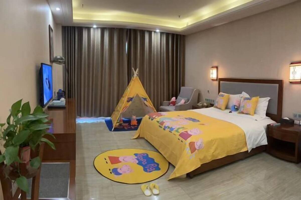 A hotel room with a bed, a TV, a desk, a chair, a window with curtains, and a small tent.
