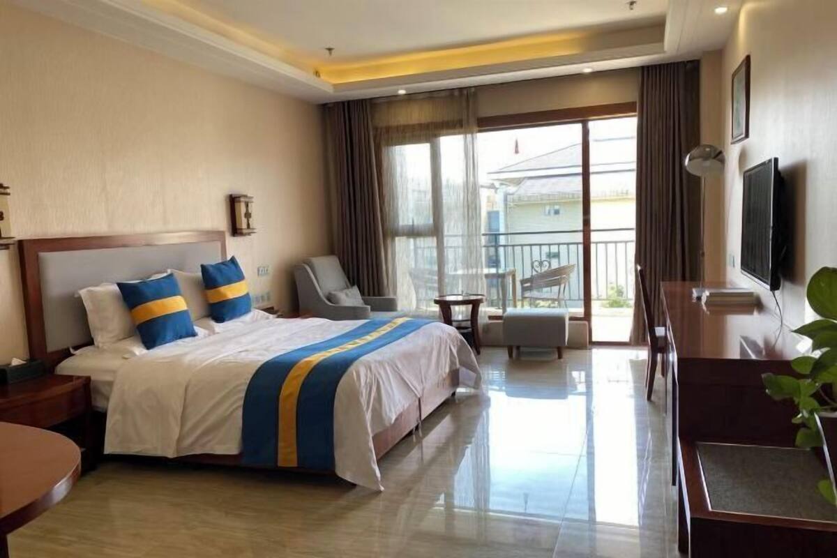 A hotel room with a large bed, a seating area with a chair and a table, a balcony with a view, and a television on the wall.