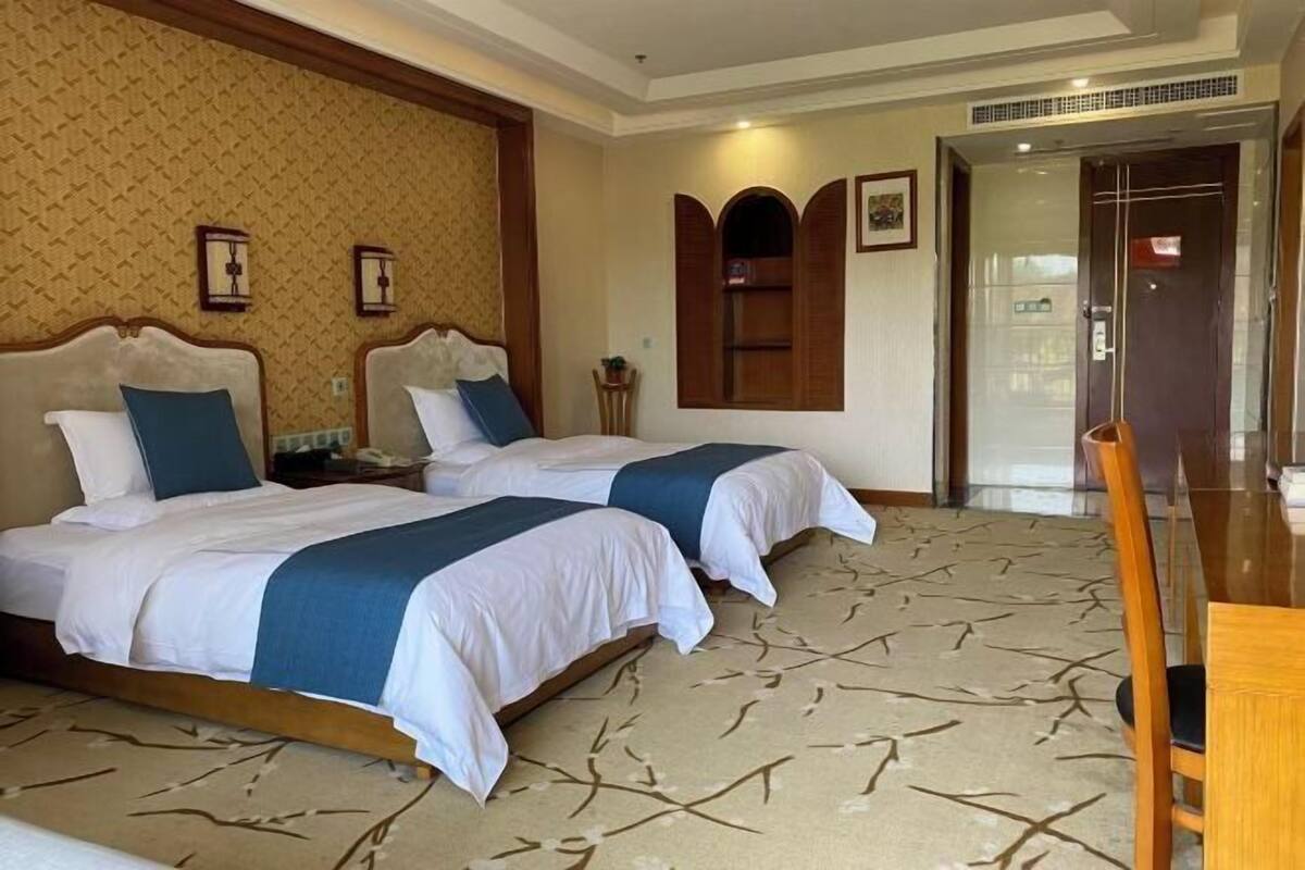 A hotel room with two beds, a wooden headboard, a patterned carpet, a wooden chair, and a door with a glass panel.