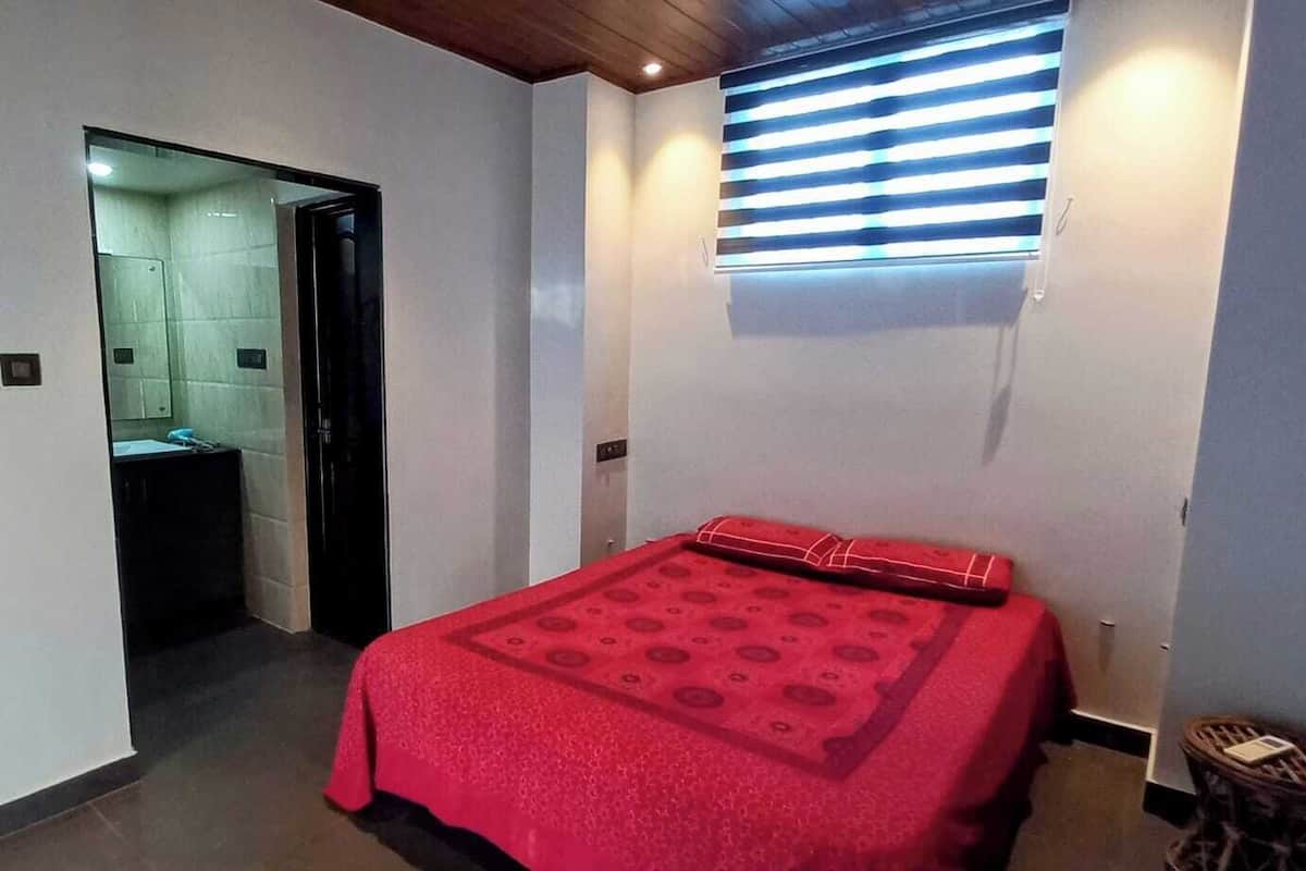 A small, narrow room with a bed covered in a red blanket, a window with horizontal blinds, and a door leading to a bathroom.