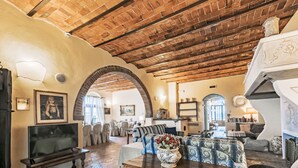 A spacious living area with a stone floor, wooden ceiling beams, and a fireplace. There is a television, seating, and a dining area visible.