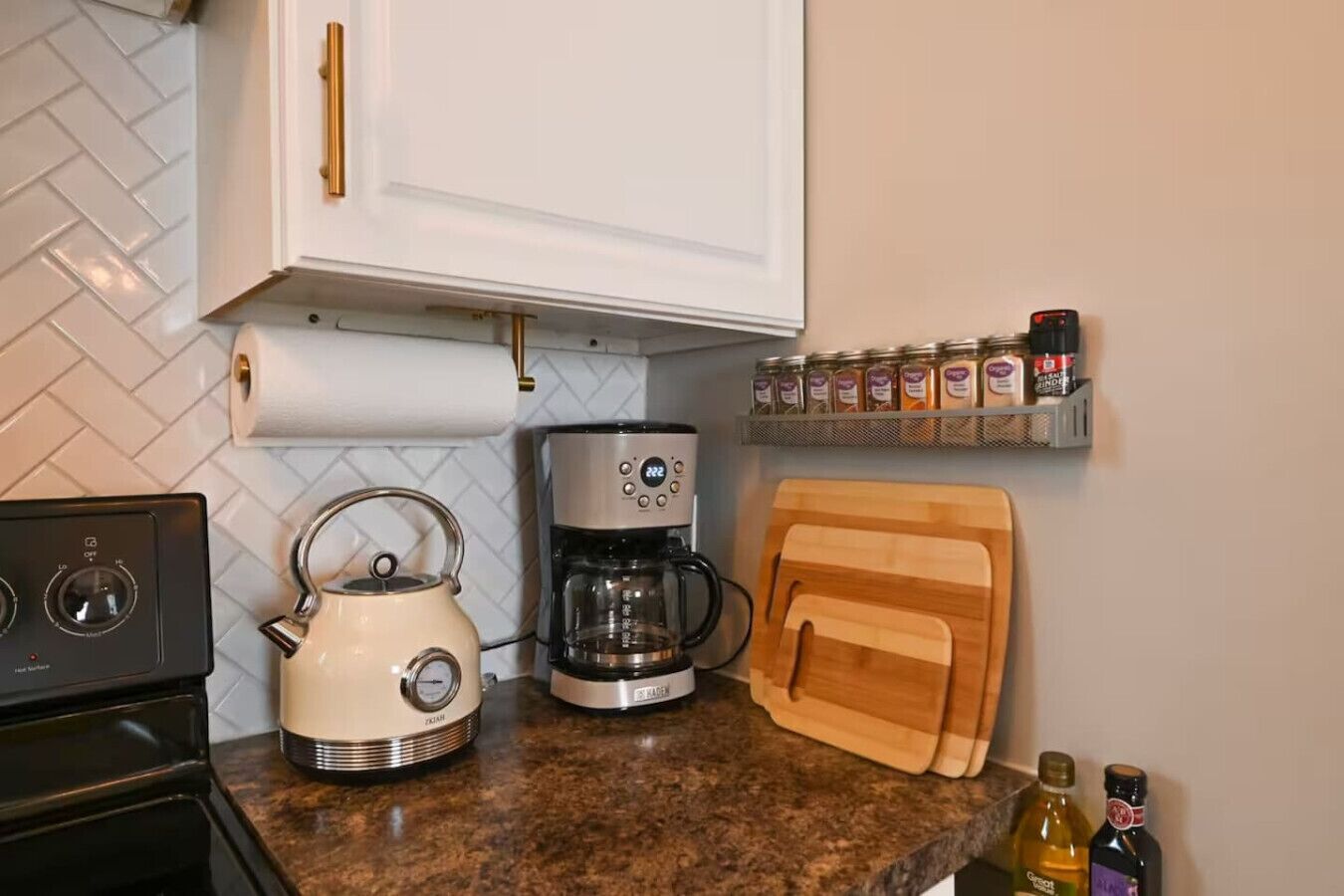 A kitchen with a stove, kettle, coffee maker, wooden cutting board, spice rack, and bottles.