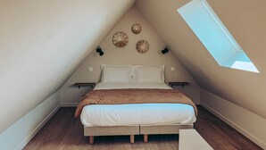 Een slaapkamer met een schuine kap, een dakraam, een bed met wit beddengoed, een bruine plaid en decoratieve muurkunst.
