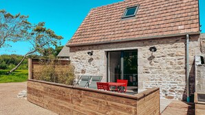 Een stenen huis met een pannendak, een houten terras en een glazen deur die leidt naar een zitgedeelte buiten met rode stoelen en een tafel.