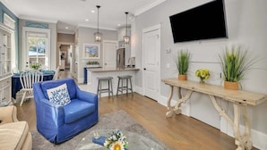 A modern living room with a blue sofa, a dining area with a table and chairs, a kitchen with a refrigerator, and a mounted TV. - 66 Palmetto Lane located in Ocean Oaks on Jekyll (Jekyll Island)