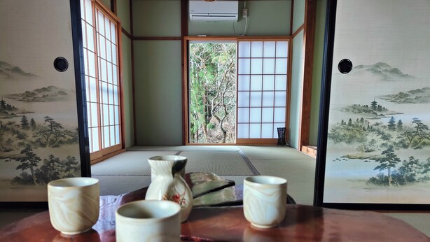 Ein traditioneller japanischer Raum mit Tatami-Boden, Shoji-Schirmen und Blick auf einen Wald durch die Schiebetüren.