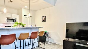 Une cuisine moderne équipée d’un four à micro-ondes, d’un évier et d’un coin bar avec des tabourets. On y trouve également une télévision et une petite table avec une plante.
