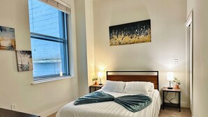 Une chambre à coucher avec un grand lit, deux tables de chevet avec des lampes, une fenêtre avec des stores et un tableau au mur.