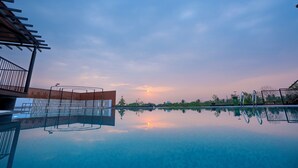 Vista despejada de una piscina con cubierta de madera y barandillas de metal, rodeada de follaje y un atardecer al fondo.