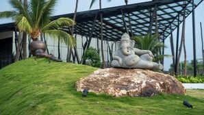 A stone statue of an elephant deity on a rock, surrounded by grass and palm trees, with a modern building in the background.