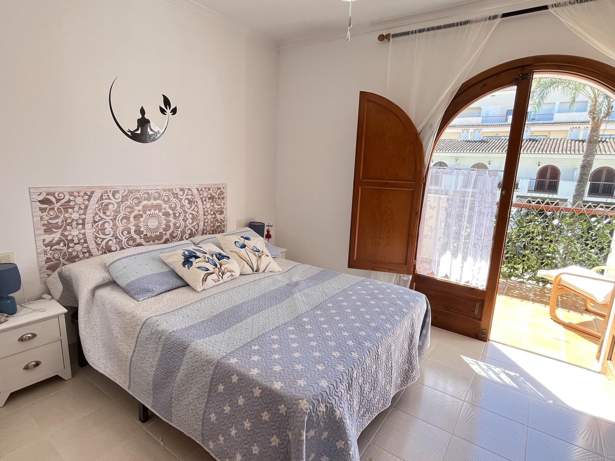 A bedroom with a bed, nightstands, a decorative headboard, a window with curtains, and a view of buildings outside.