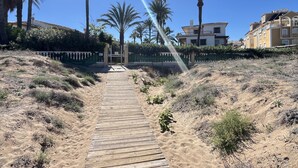 A wooden pathway leading to a gated entrance, surrounded by dry grass and palm trees, with buildings in the background. - Duplex apartament Bahia Azul on the beachfront for 4 persons (Dénia)