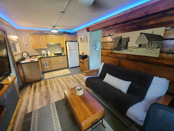 A compact living space with a kitchenette, a wooden dining table, a black and white sofa, and a rustic wall with a painting.