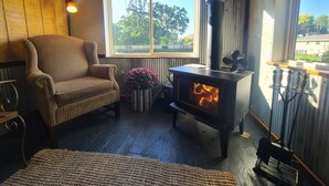 Une pièce chaleureuse avec un poêle à bois, un fauteuil confortable, une fenêtre donnant sur les arbres et une lampe fixée au mur.