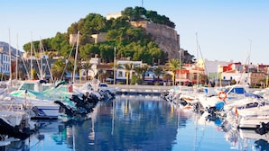 A marina with boats docked in calm waters, a historic castle on a hill in the background, and buildings along the waterfront. - Essence by Interhome (Dénia)