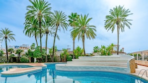 A swimming pool with a clear blue water feature, surrounded by palm trees and a view of buildings in the background. - Essence by Interhome (Dénia)