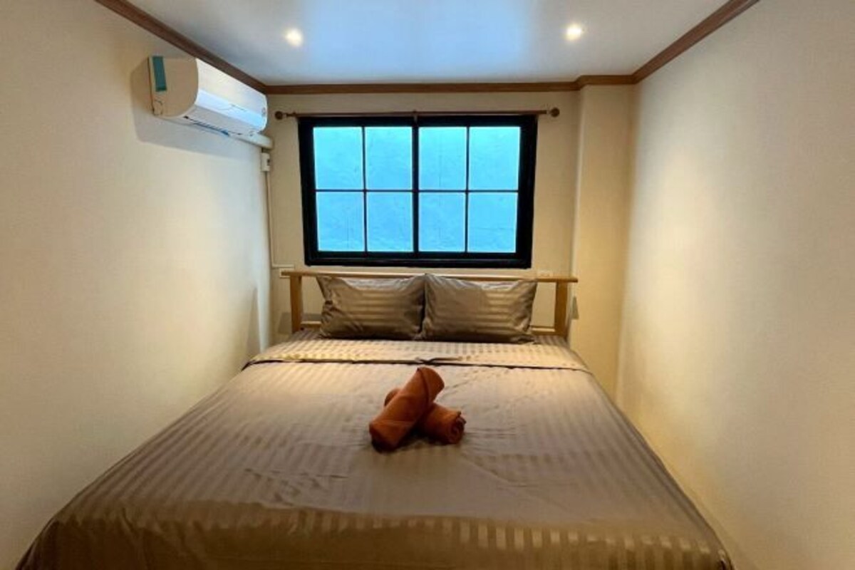 A small, neatly made bed with a window above it, a wall-mounted air conditioner, and a simple, clean room layout.