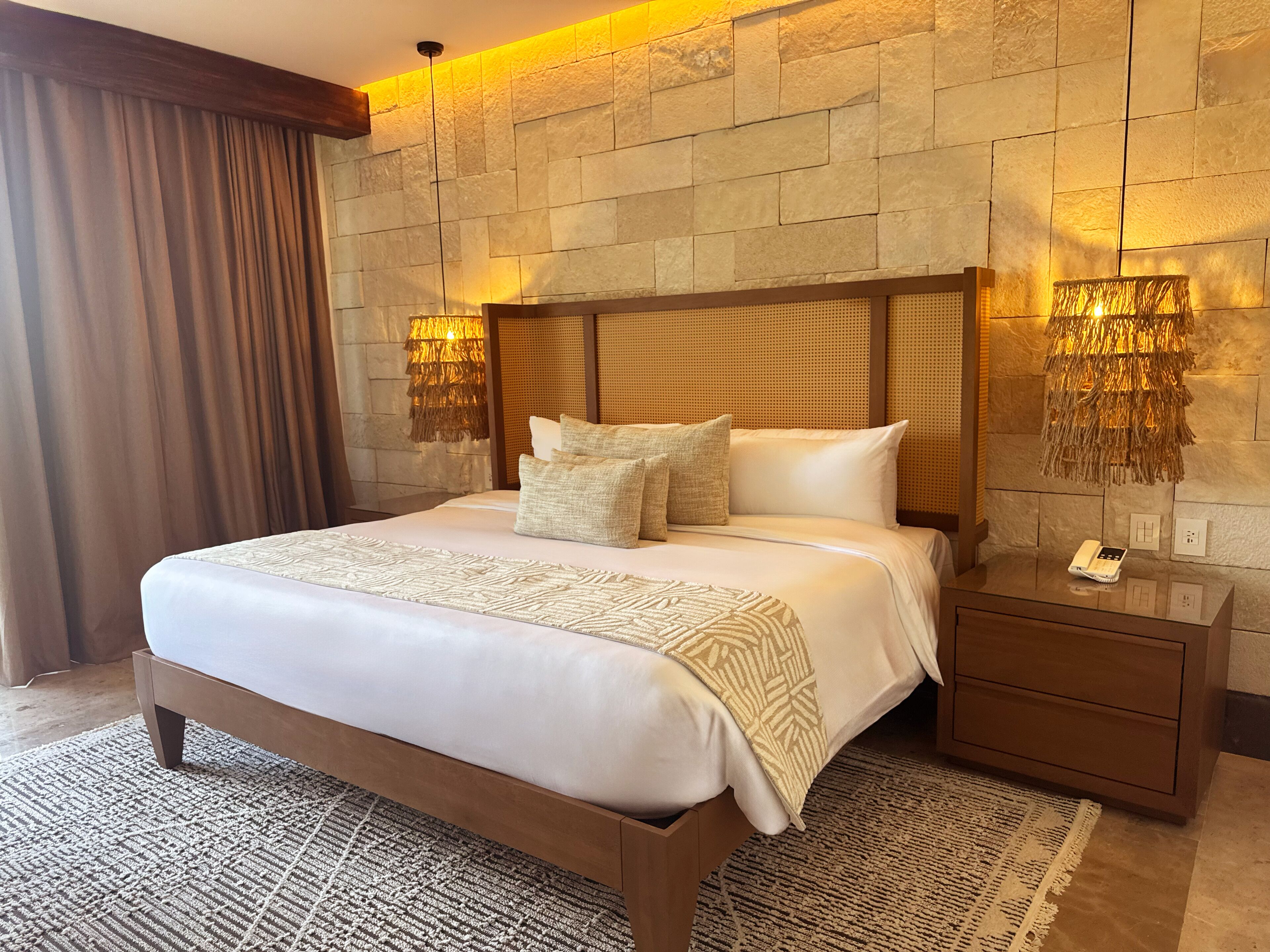 A hotel room with a large bed, wooden headboard, two bedside lamps, a nightstand, and a textured rug.