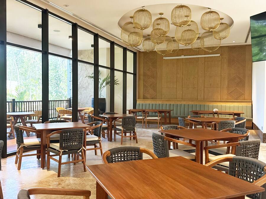 Uma ampla sala de jantar coberta, com mesas e cadeiras de madeira, janelas grandes e um lustre decorativo.