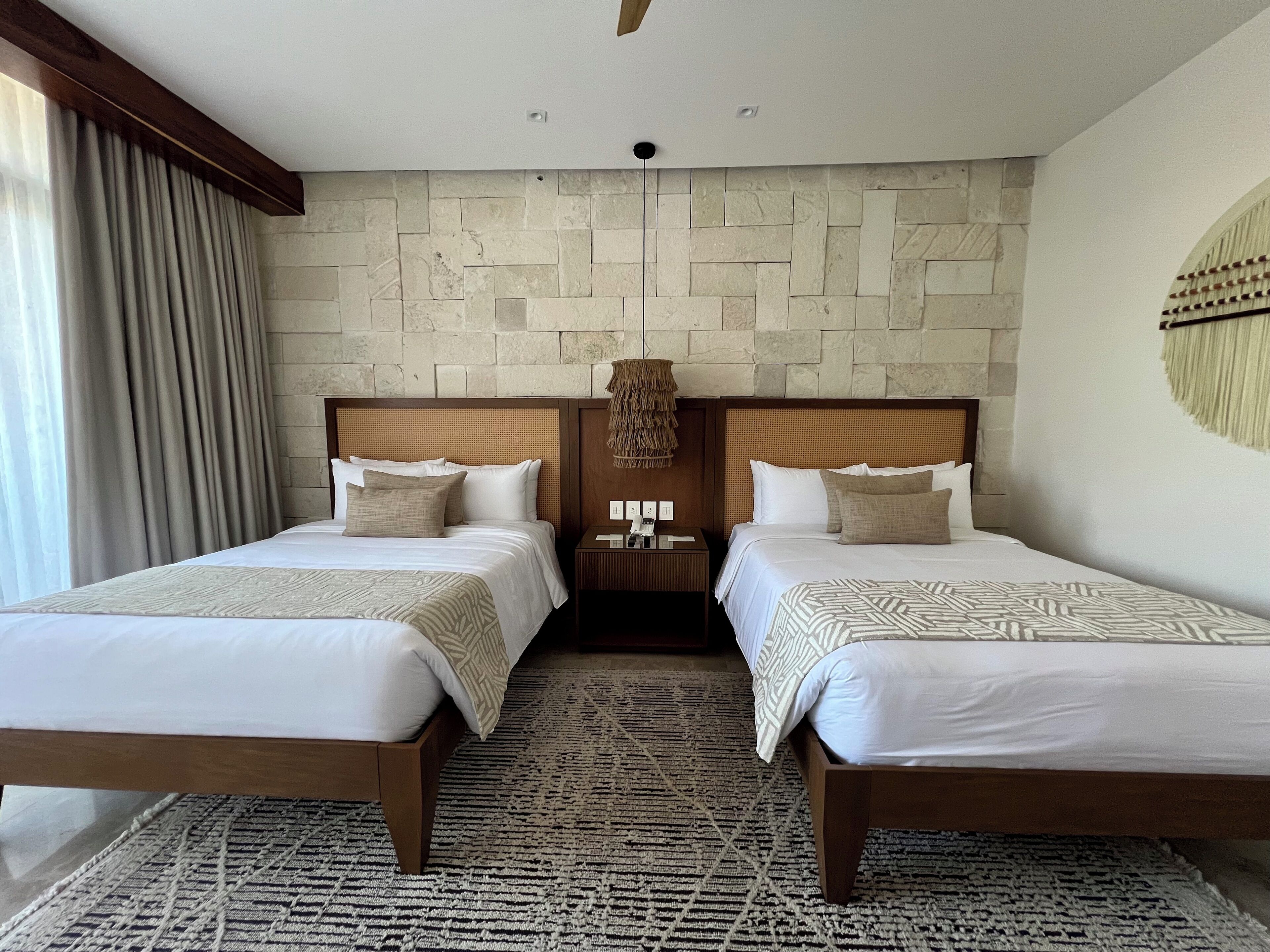 Une chambre d’hôtel avec deux lits, un mur en pierre, une tête de lit en bois, un tapis à motifs et une table de chevet avec une lampe.