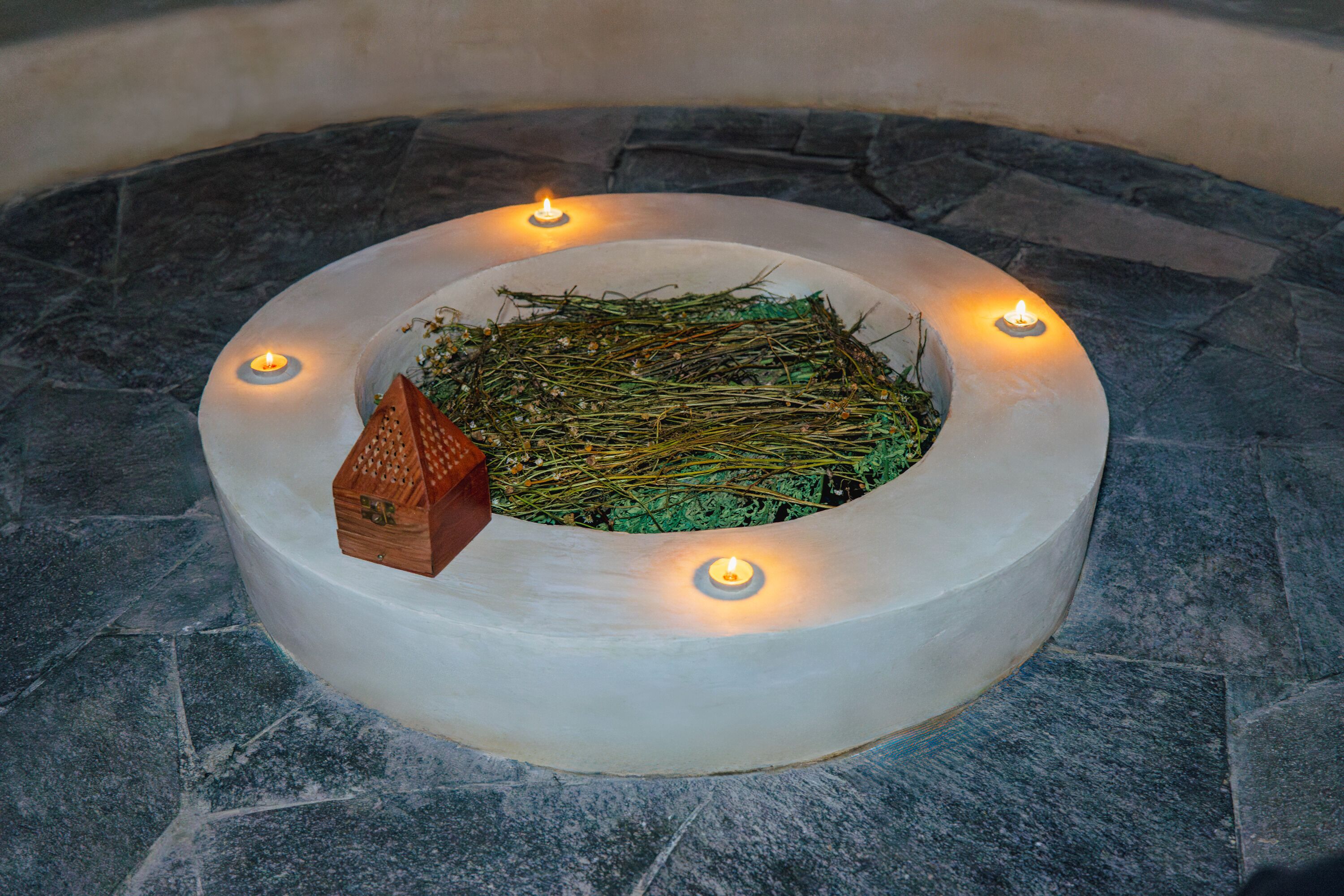 Una estructura de piedra circular con un montón central de ramas y un pequeño objeto de madera, rodeada por cuatro velas encendidas.