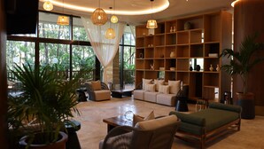 A spacious living area with a sofa, chairs, and a coffee table. There are large windows with curtains, a bookshelf, and potted plants. - Hotel Mundo Maya Tulum (Tulum)