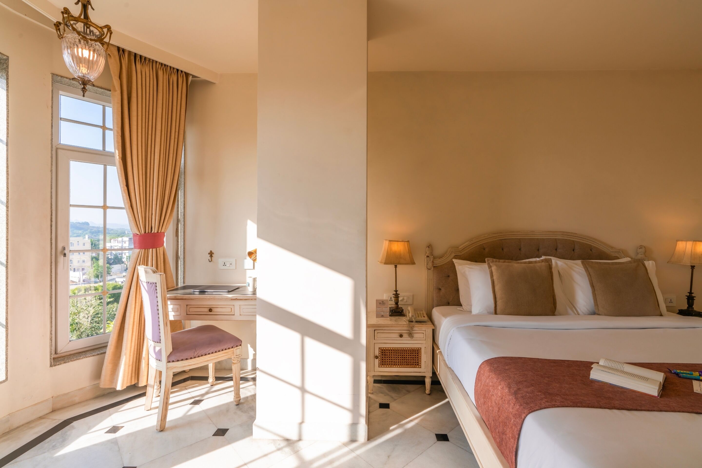 A hotel room with a bed, bedside table, lamp, chair, desk, and a window with curtains.
