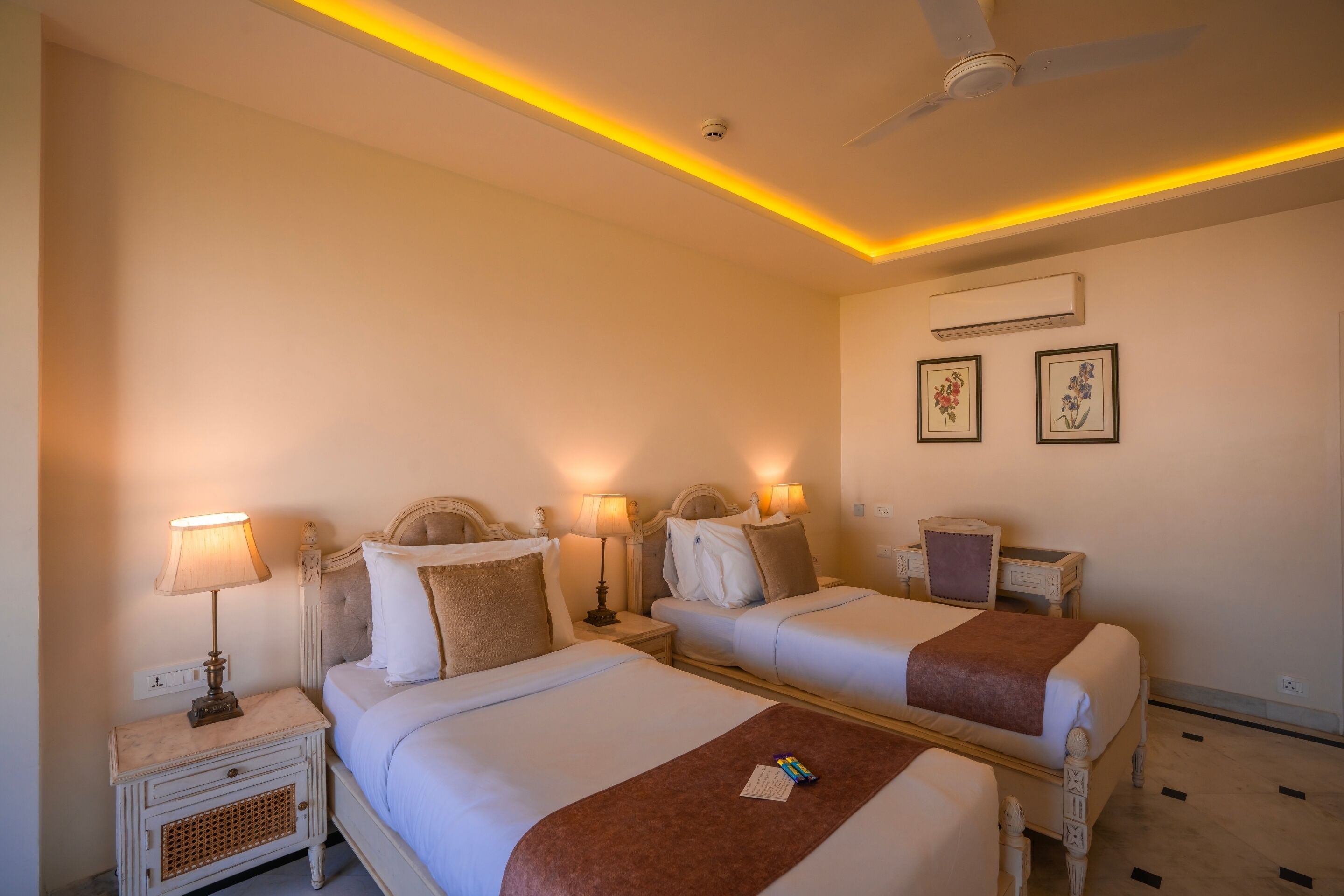 Habitación de hotel con dos camas, mesitas de noche con lámparas, ventilador de techo y cuadros en la pared.