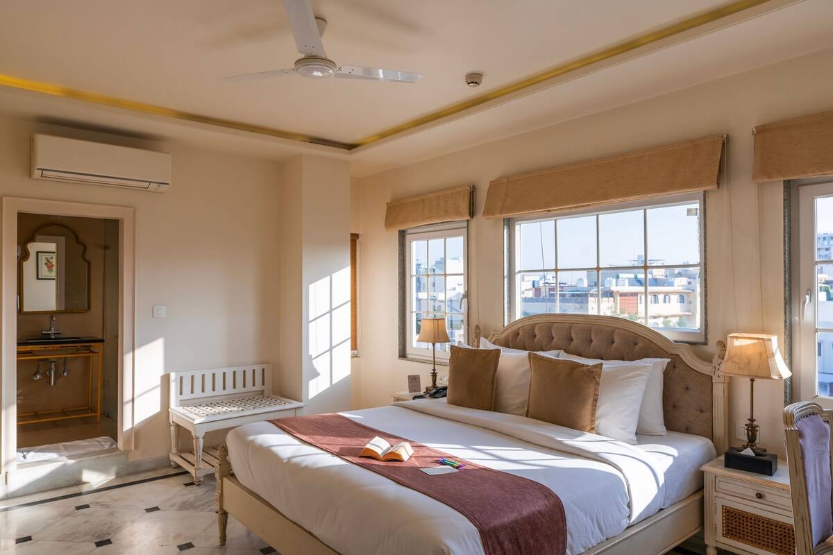 A hotel room with a bed, bedside tables, a window with a view of buildings, an air conditioning unit, and a ceiling fan.