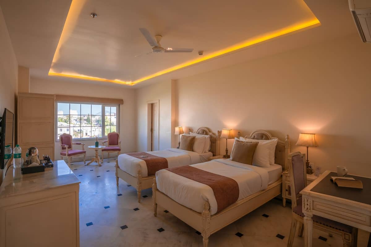 A hotel room with two beds, a desk, a chair, a small table, and a window with a view of buildings.