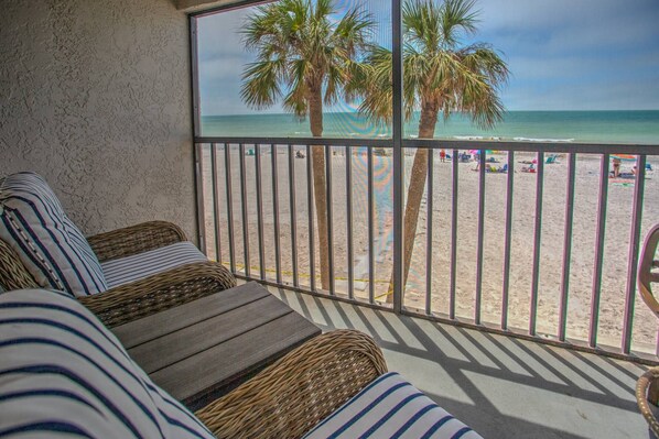 Un balcone con mobili in vimini che si affaccia su una spiaggia con palme e persone.