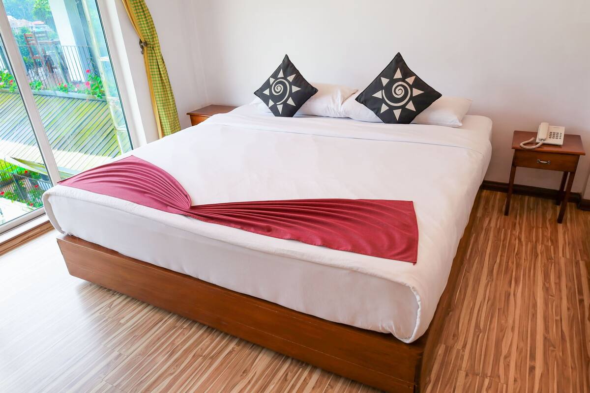 A modern hotel room with a large bed, a wooden floor, a balcony with a view, and a bedside telephone.