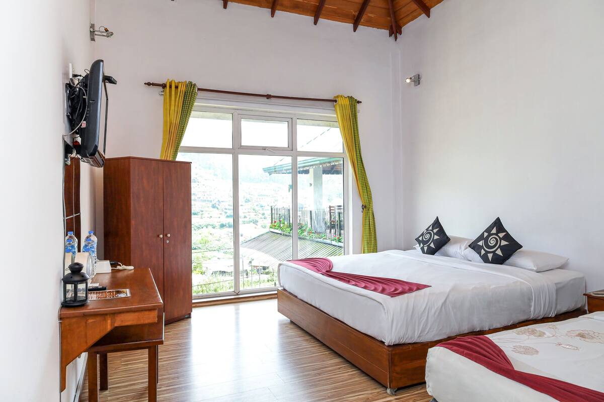 A bedroom with a large window, a wooden wardrobe, a desk with a lamp, and a bed with pillows.