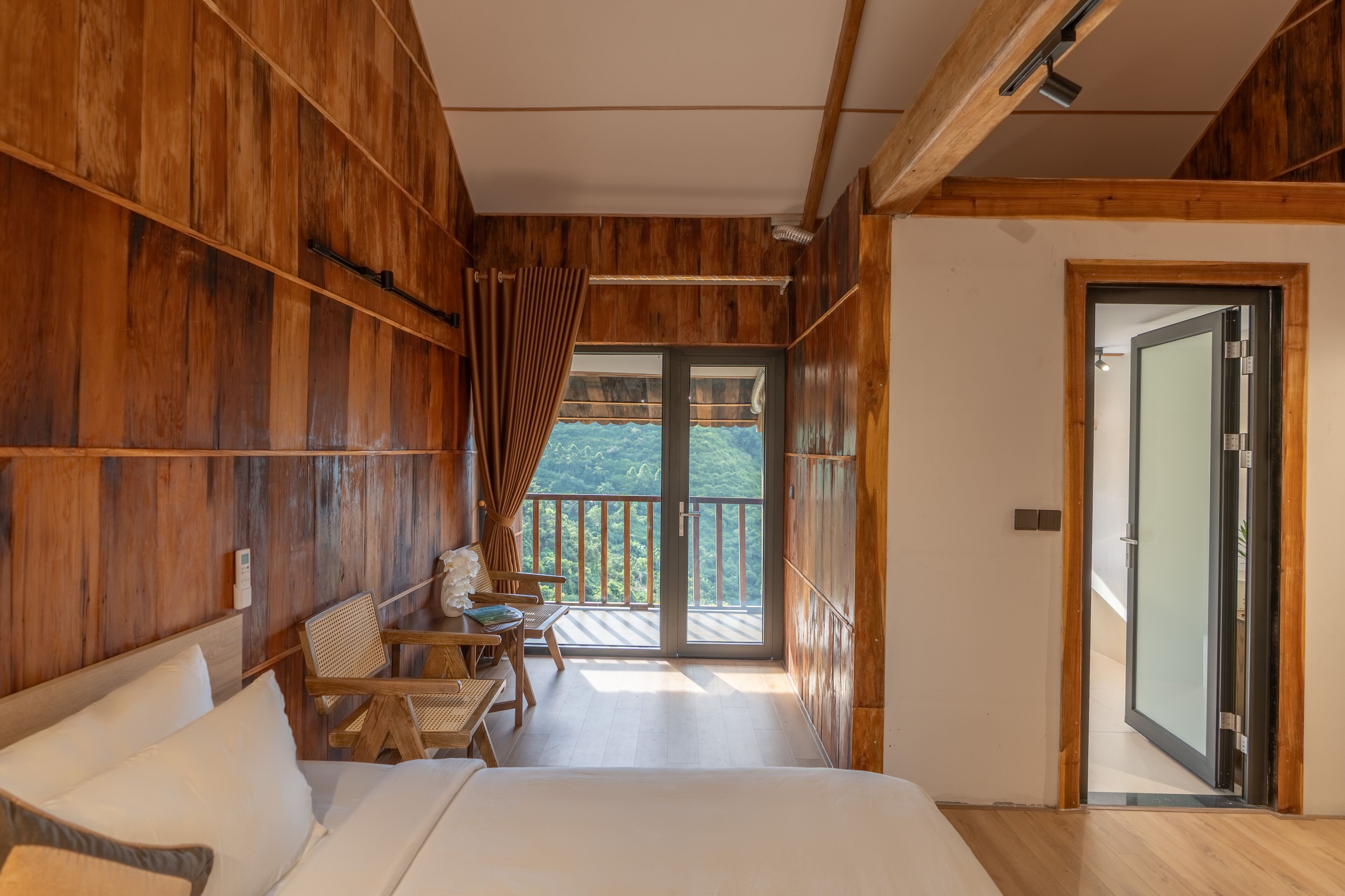 A wooden room with a bed, a table, and chairs. There is a balcony with a view of the outdoors.