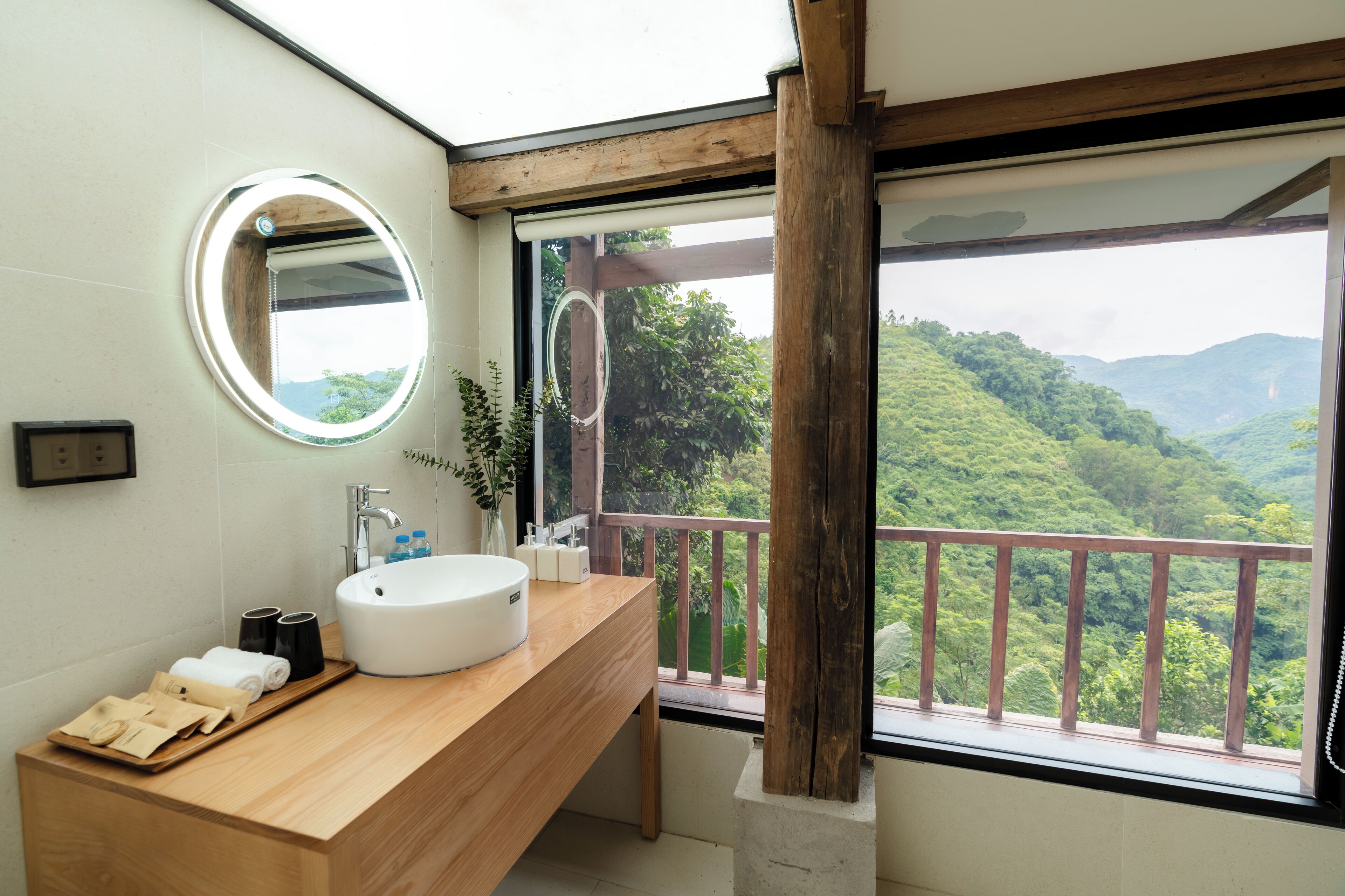 A bathroom with a wooden vanity, a round mirror, a white sink, and a view of a lush green landscape through a large window.