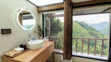A bathroom with a wooden vanity, a round mirror, a white sink, and a view of a lush green landscape through a large window.