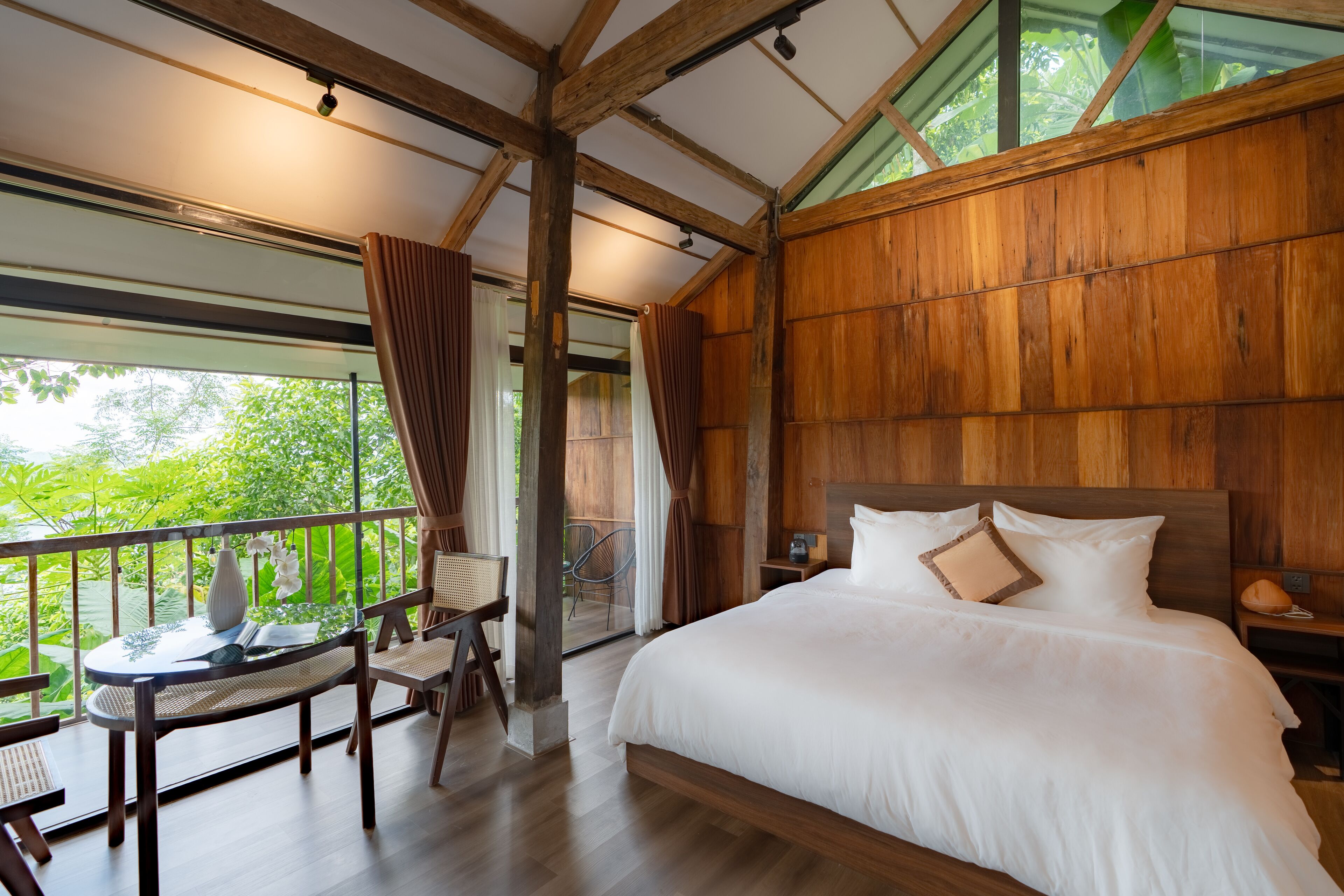 Een slaapkamer met een groot bed, houten wanden, een balkon met tafel en stoelen, en uitzicht op groen.