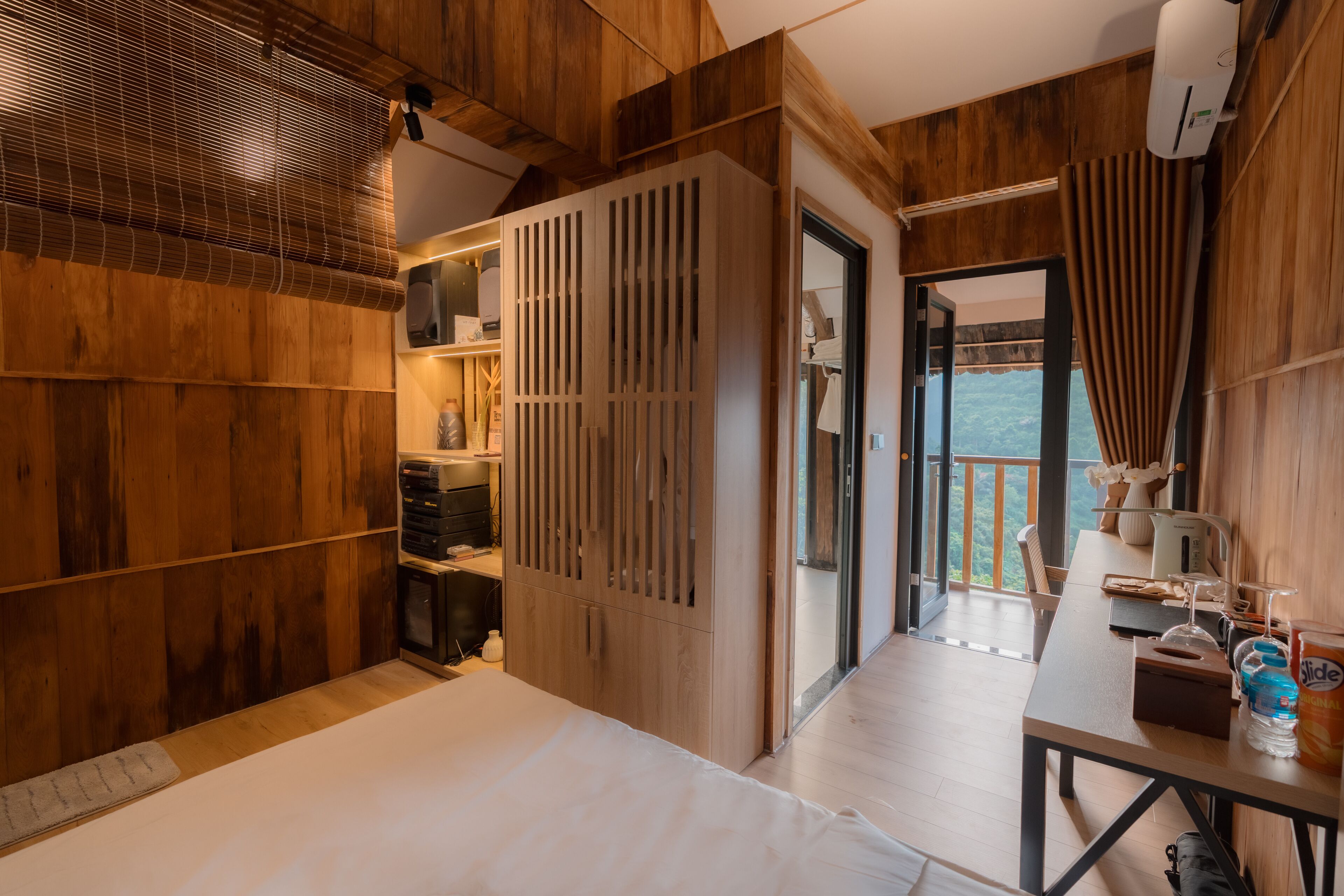 Quarto de madeira com uma cama, uma mesa com objetos e vista para o exterior.